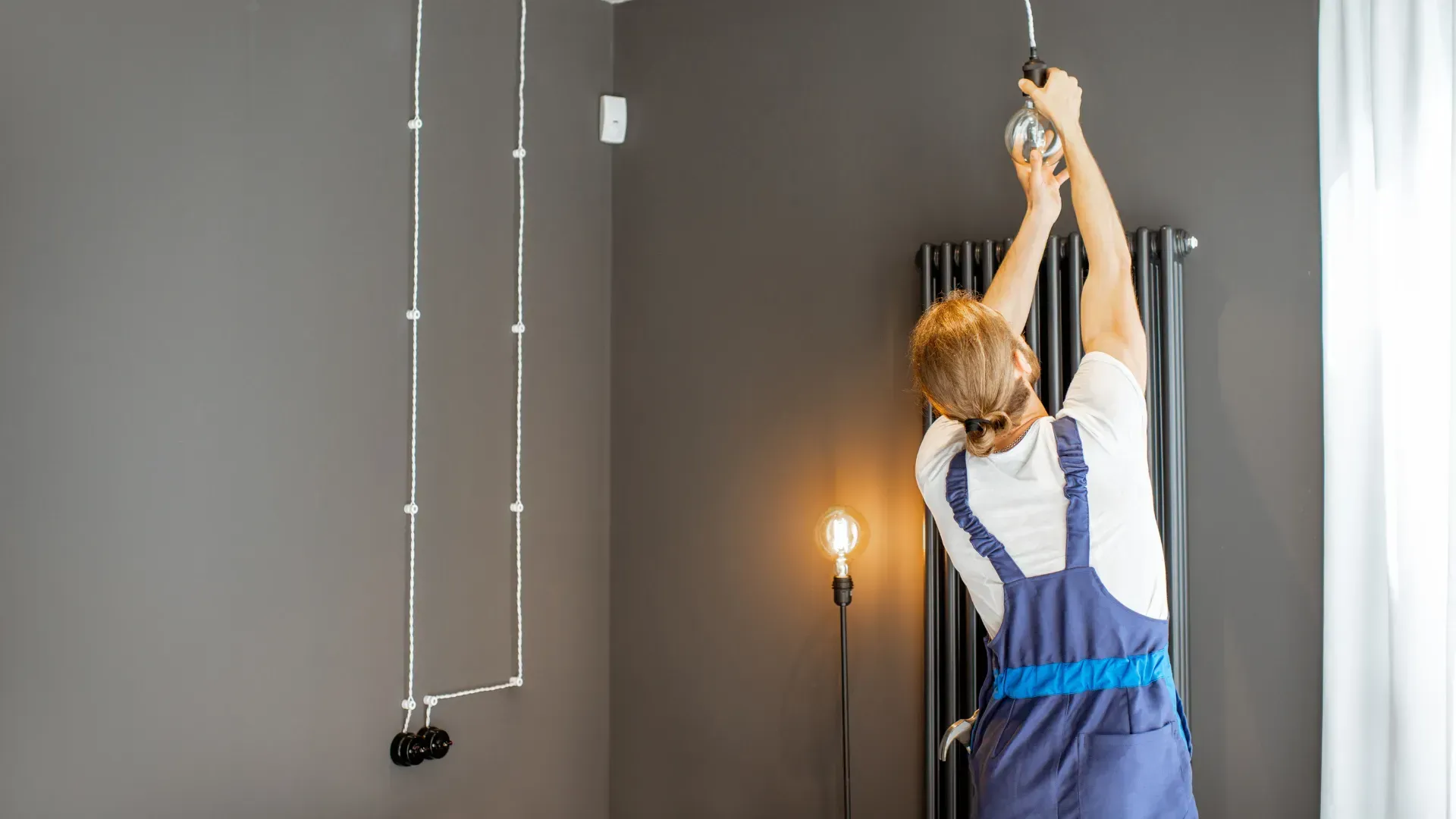 Man in overalls installing light fixture near a grey wall.