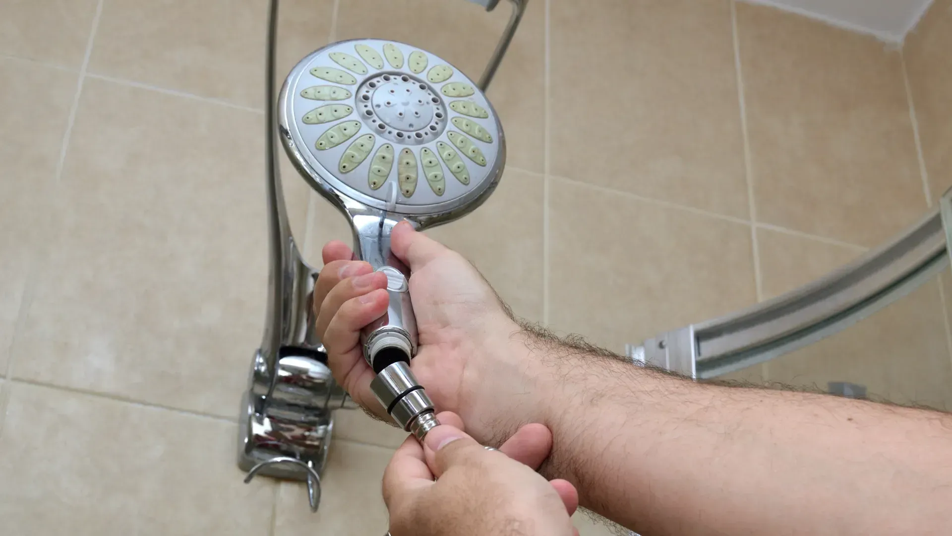 Person's hands unscrewing a showerhead from the hose in a tiled shower.