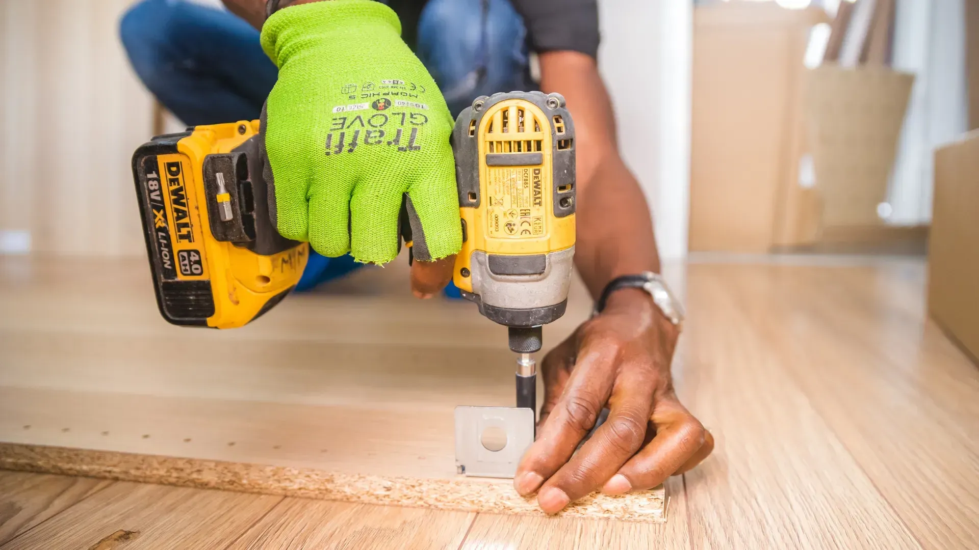 Person assembling furniture with a power drill, wearing green gloves, on a wood floor.