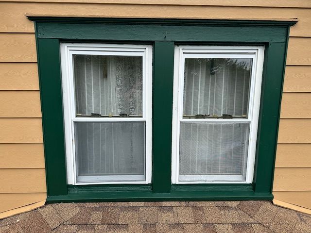 Two white-framed windows with green trim in a yellow siding wall. Dark green trim.