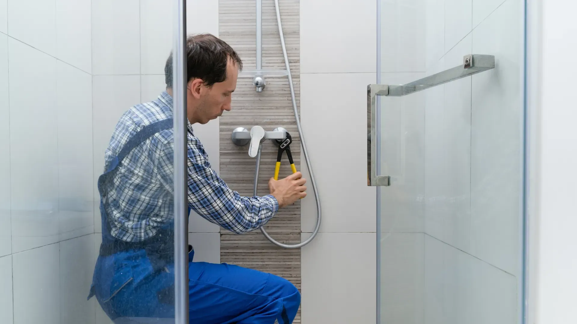 Man in blue overalls, using pliers on showerhead in a tiled shower with glass door.