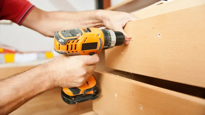 Hands using an orange drill to fasten something to a wooden structure.