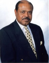 Man in a suit and tie, smiling, looking at the camera. He has a mustache and a patterned tie.