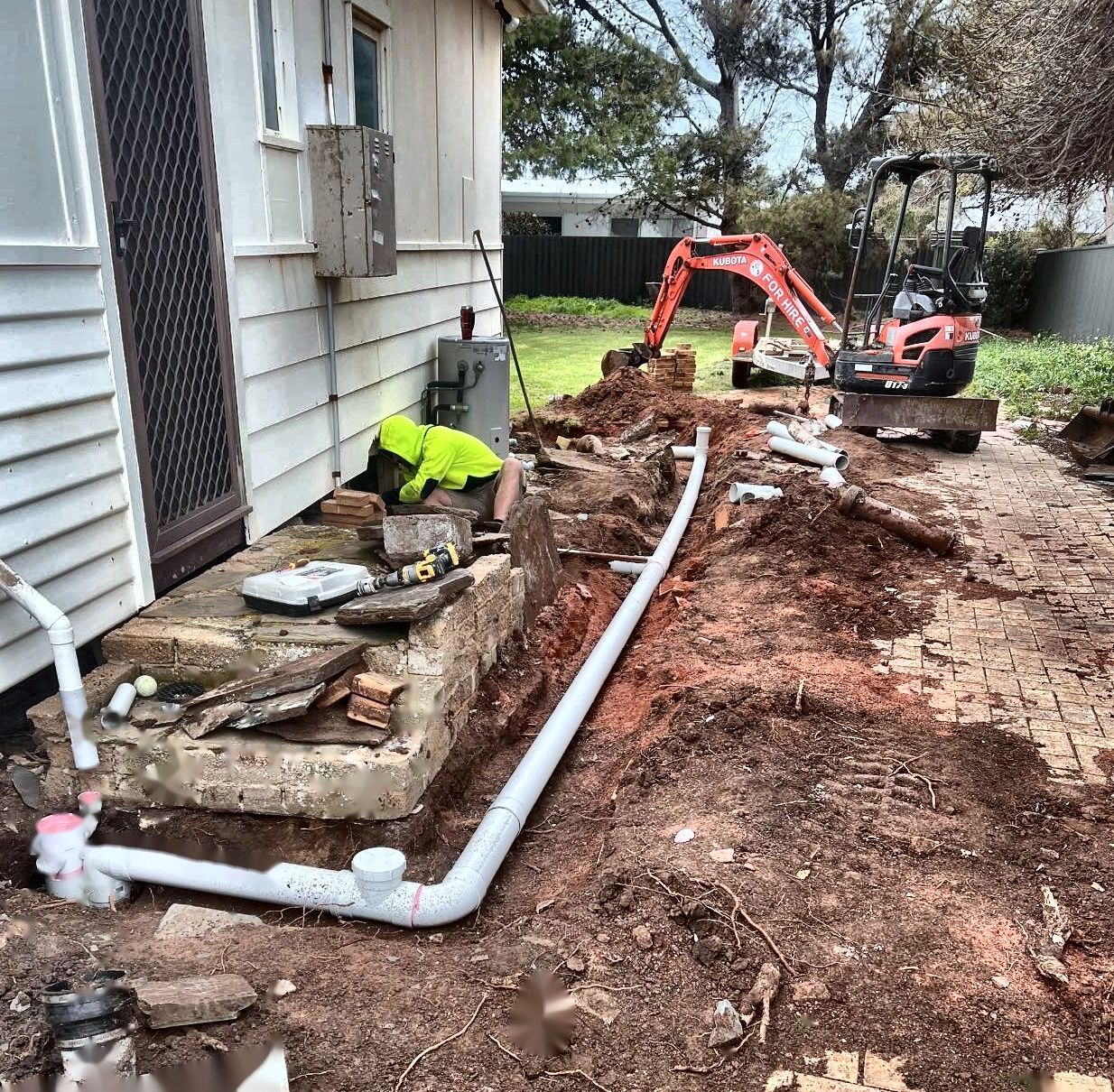 Construction site: White pipes in a trench next to a house with an excavator in the background. A worker in yellow vest.