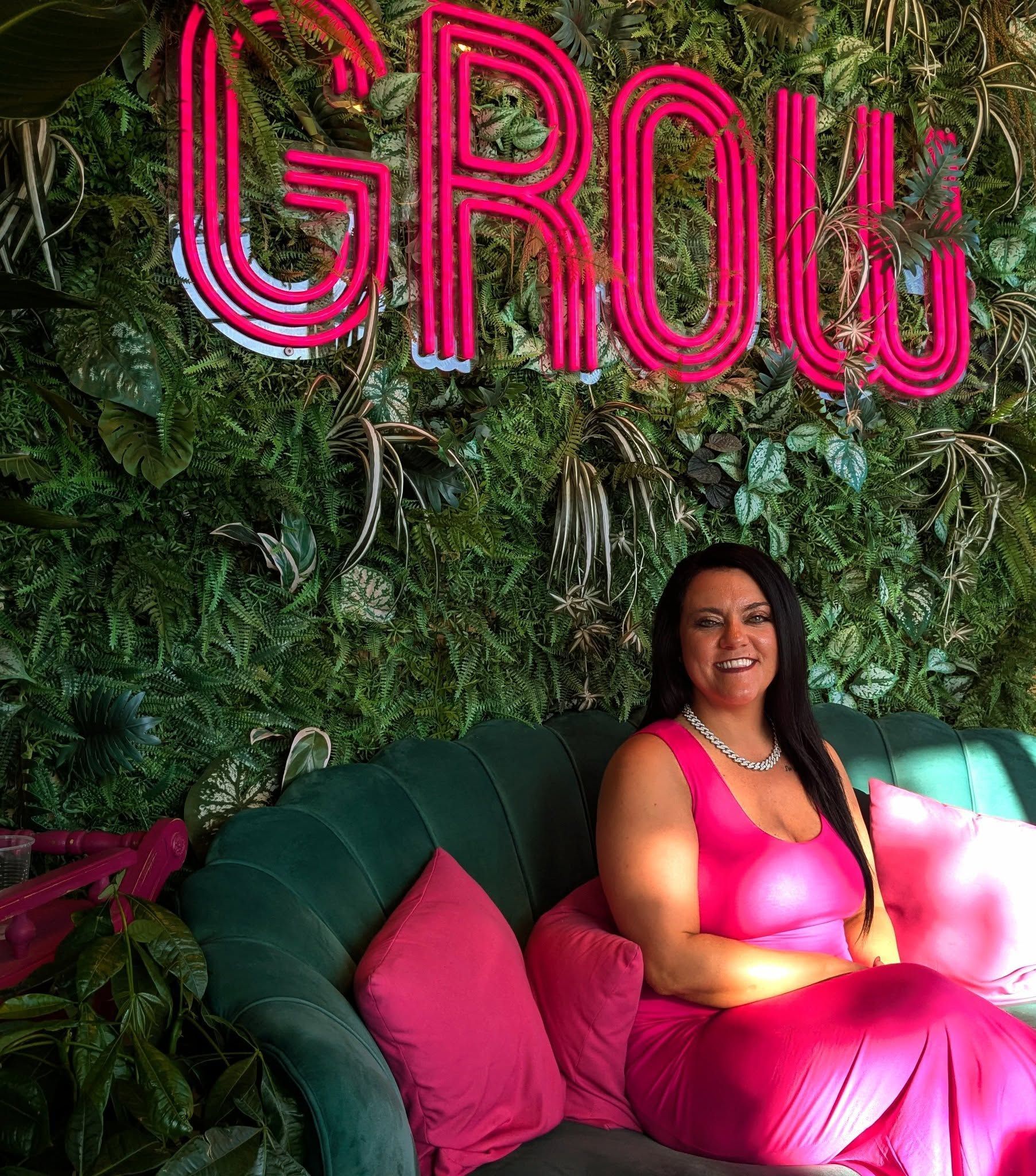 Woman in a bright pink dress sitting on a green couch before a lush wall with neon pink GROW sign