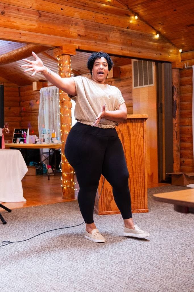 Person dancing in a rustic wooden room, one arm raised, wearing a beige top, black pants, and white sneakers.