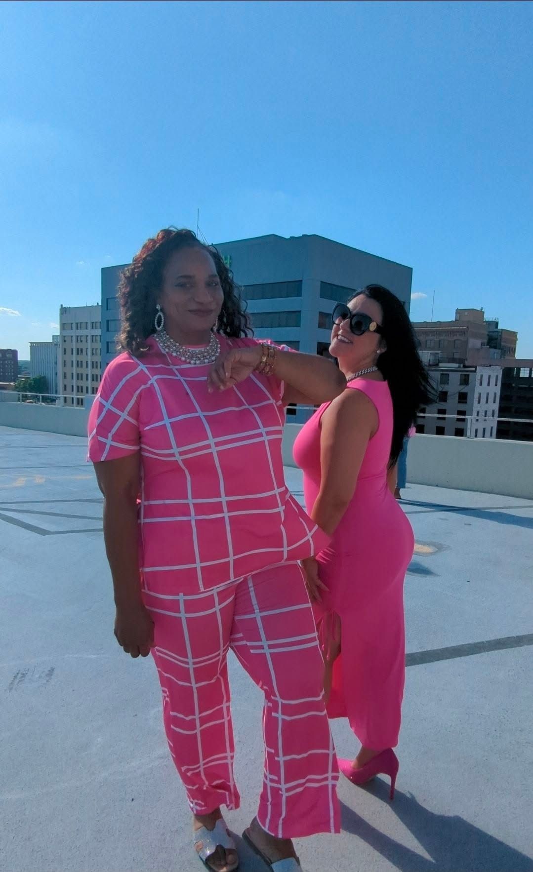 Two people in bright pink outfits pose on a rooftop with city buildings in the background.