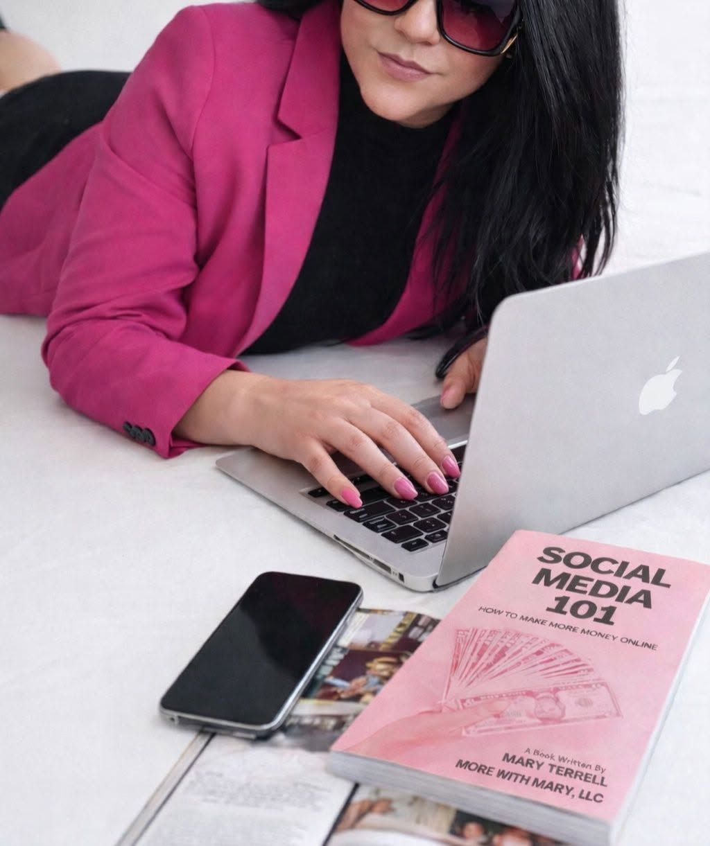 Woman in a magenta blazer using a laptop beside a phone and a pink social media book