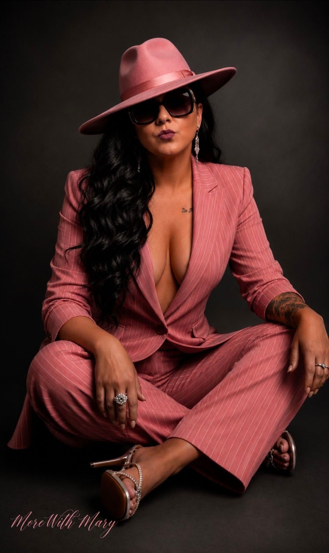 Woman in a pink suit and wide-brim hat, seated against a dark background, wearing sunglasses.