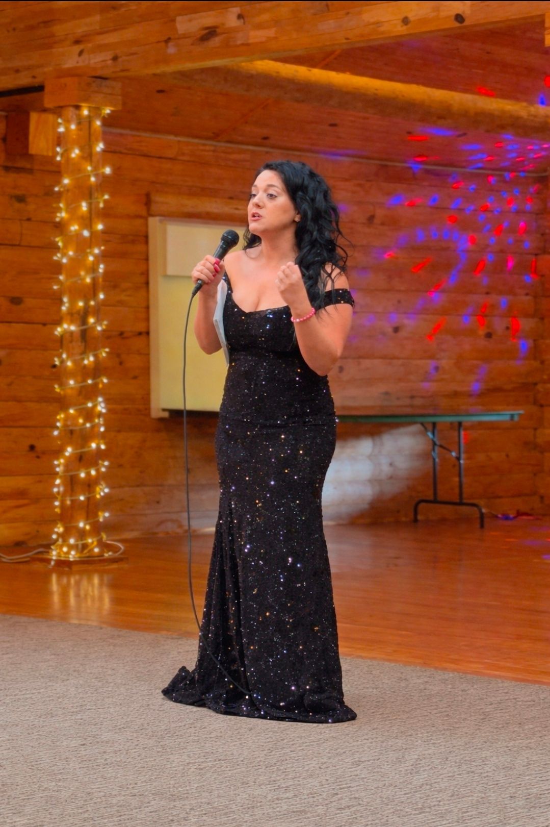 Woman in a black sequined gown singing into a microphone on a wooden stage with colorful lights