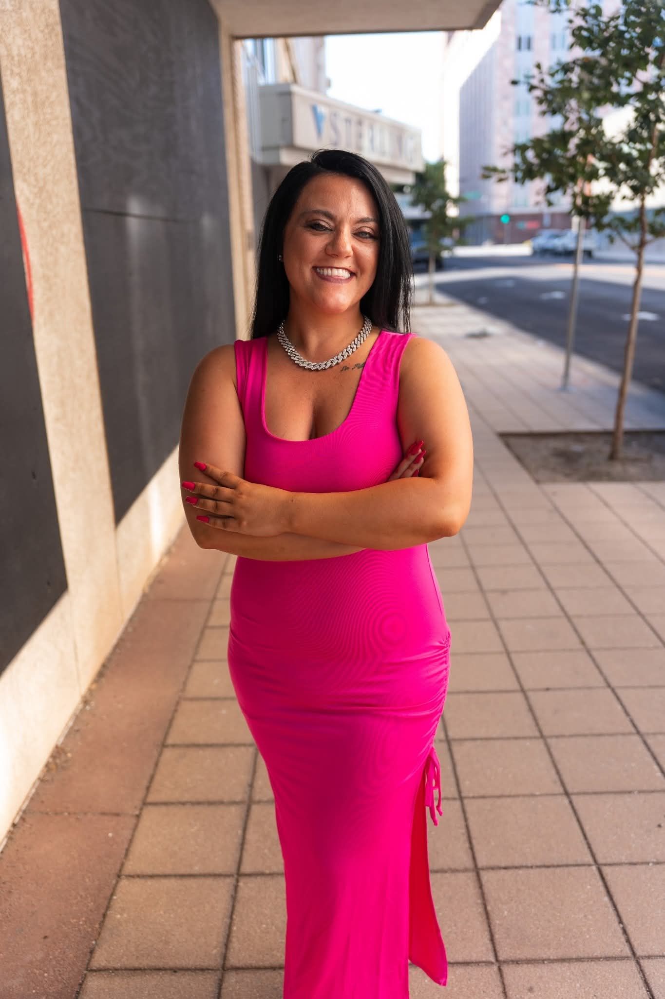 Woman in a bright pink dress smiling on a sunlit sidewalk, arms crossed.