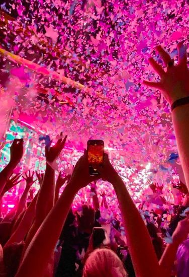 Crowd at a concert with hands raised as pink confetti falls under neon light