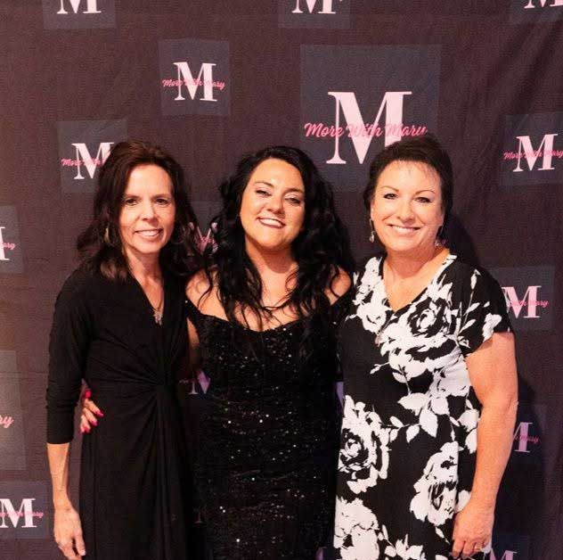 Three women posing together at a branded event backdrop, smiling in black and white outfits.