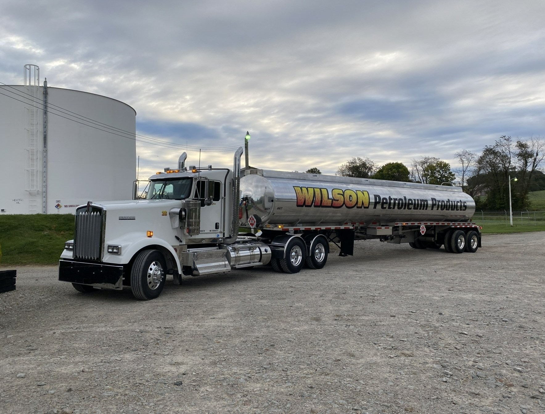A white Kenworth semi-truck with a silver tanker trailer parked on a gravel lot next to a large white fuel storage tank.