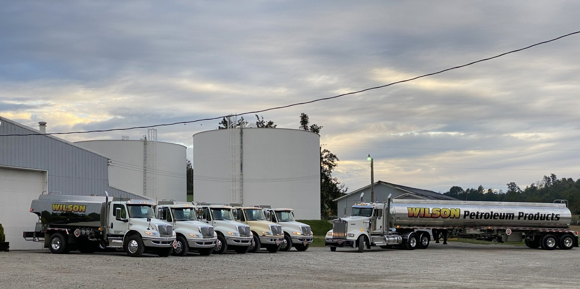 A row of several parked silver tanker trucks at an industrial site with large storage tanks under a cloudy sky.