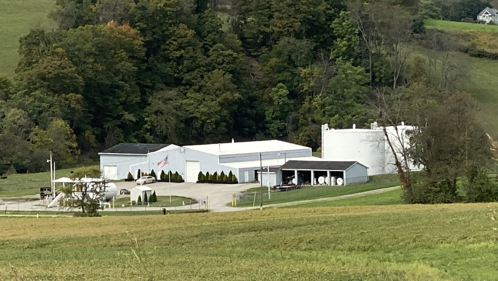 A white industrial building with large storage silos, situated in a green rural field near a treeline.