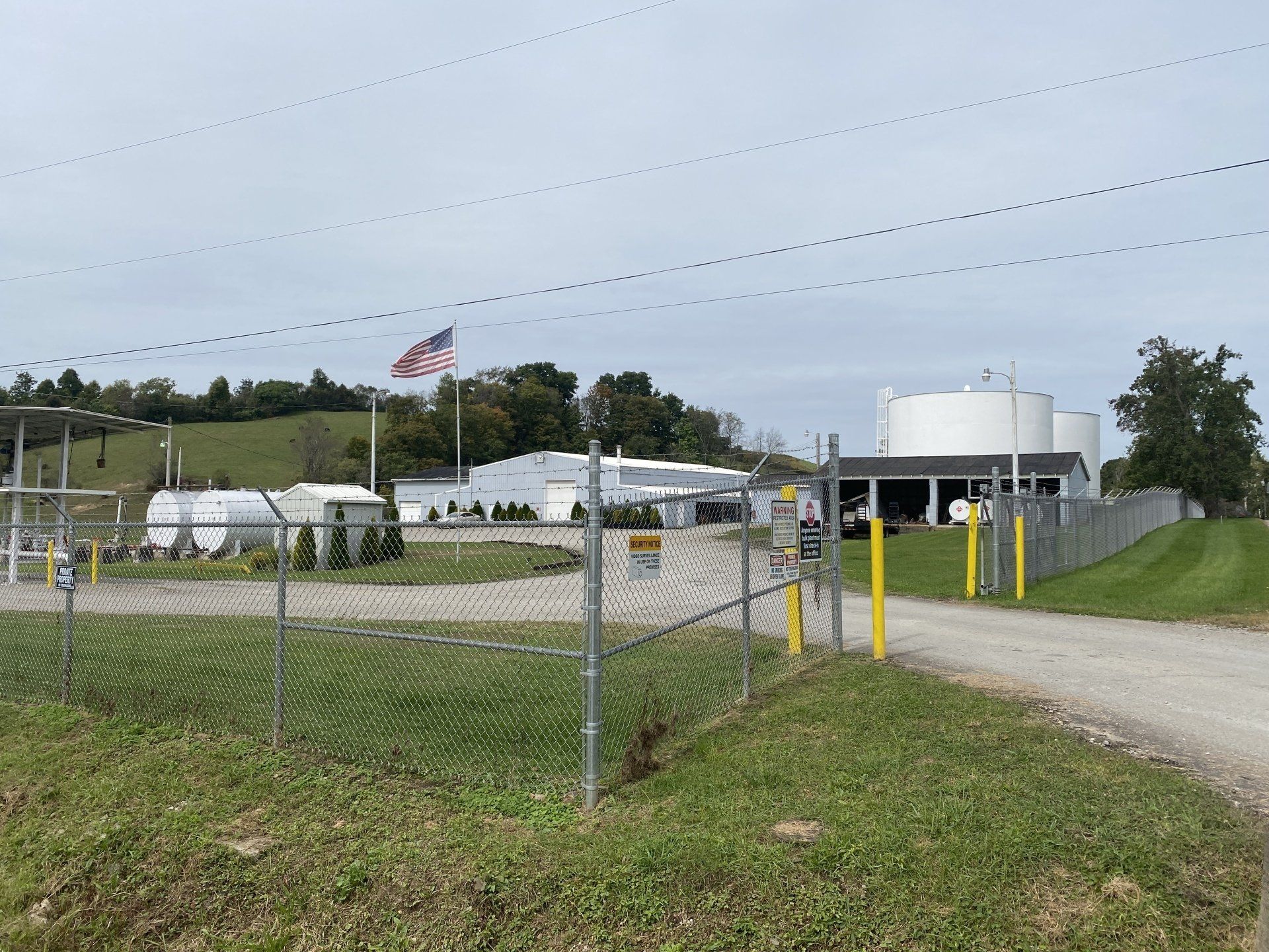 Fenced industrial facility with large white storage tanks, an American flag, and a gravel driveway under a cloudy sky.