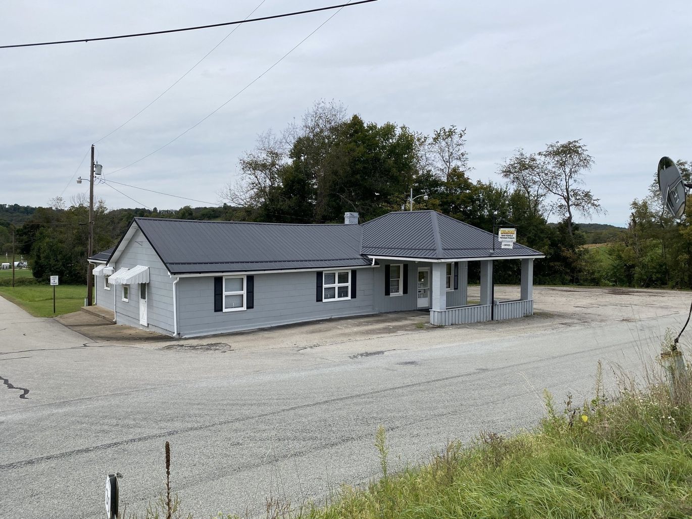 A single-story gray building with a dark roof and a gravel lot under an overcast sky in a rural setting.