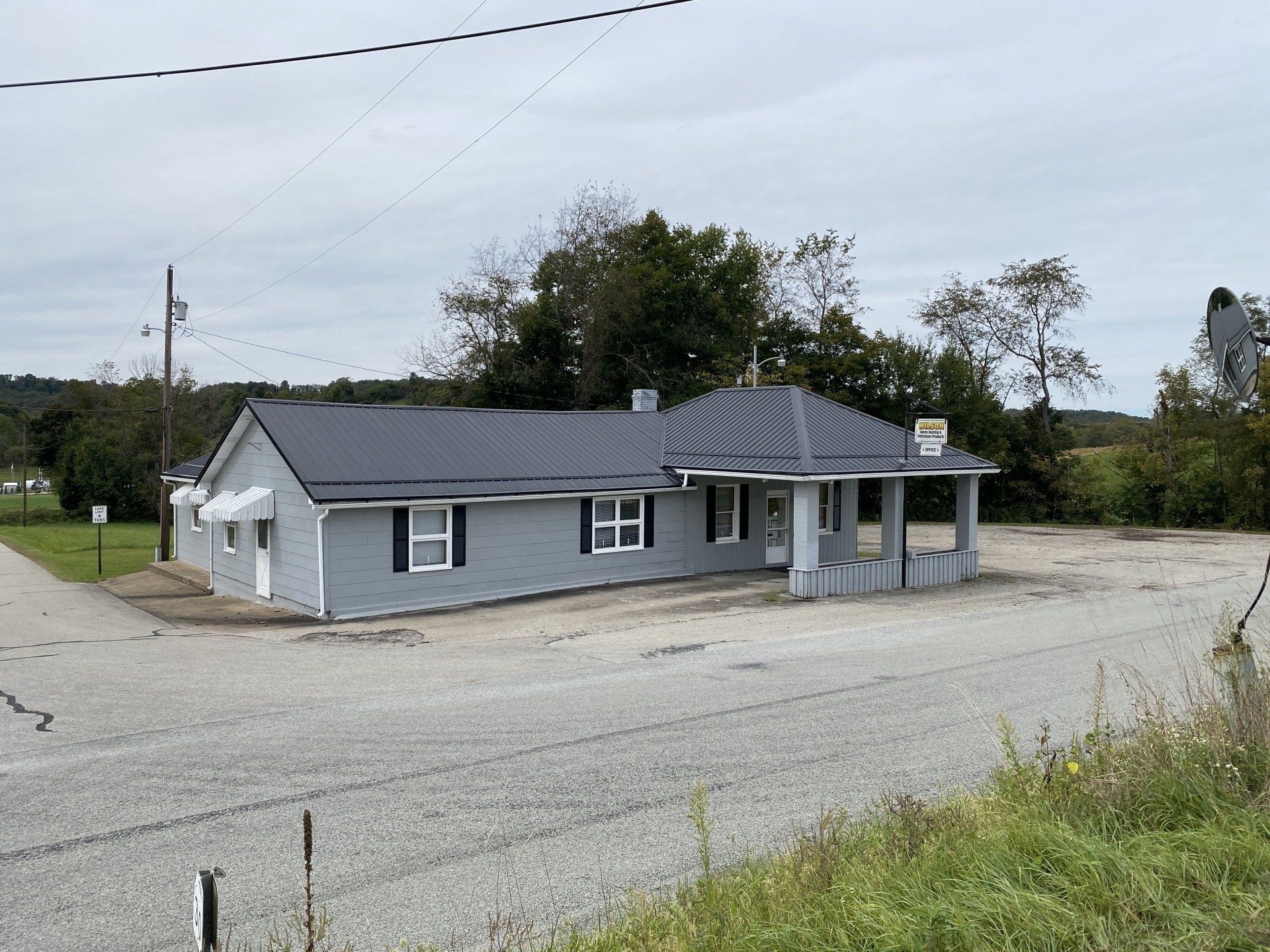 A single-story gray building with a dark roof and a gravel lot under an overcast sky in a rural setting.