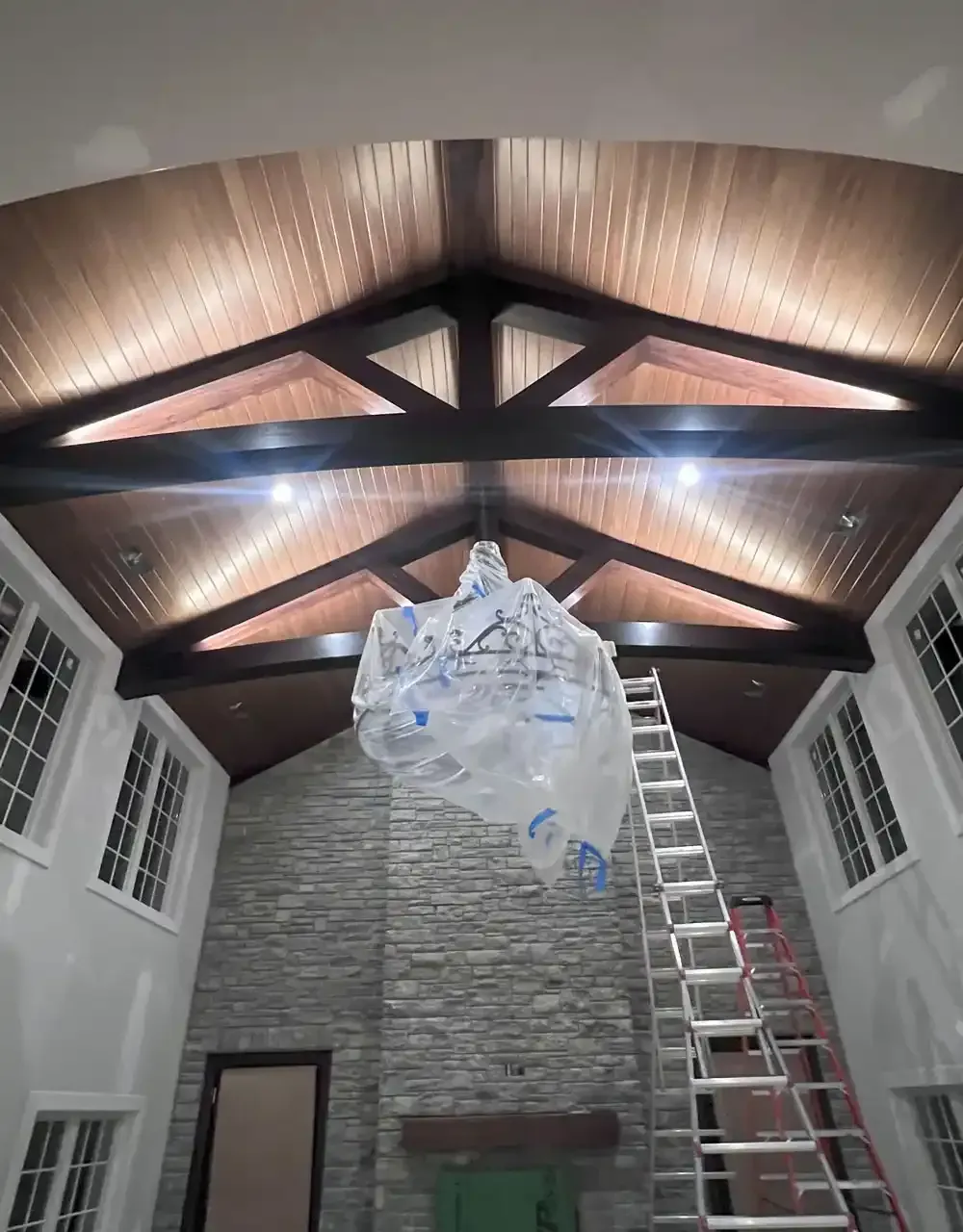 High-ceiling room with wood beams, wood paneling, and stone fireplace; chandelier wrapped in plastic, ladder present.