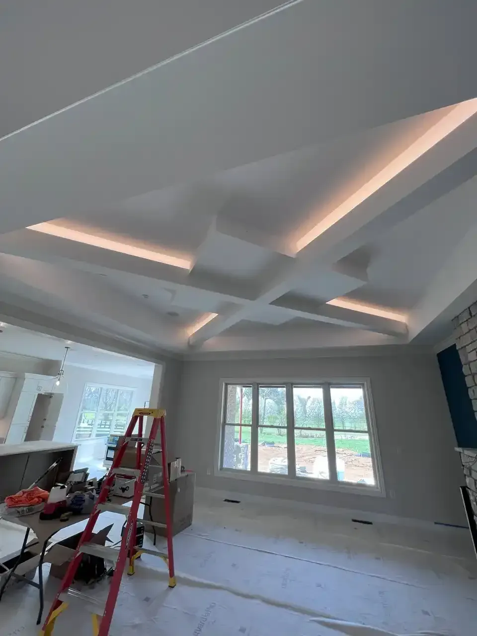 Ceiling with recessed lighting, ladder in a room under construction with a window and a fireplace.