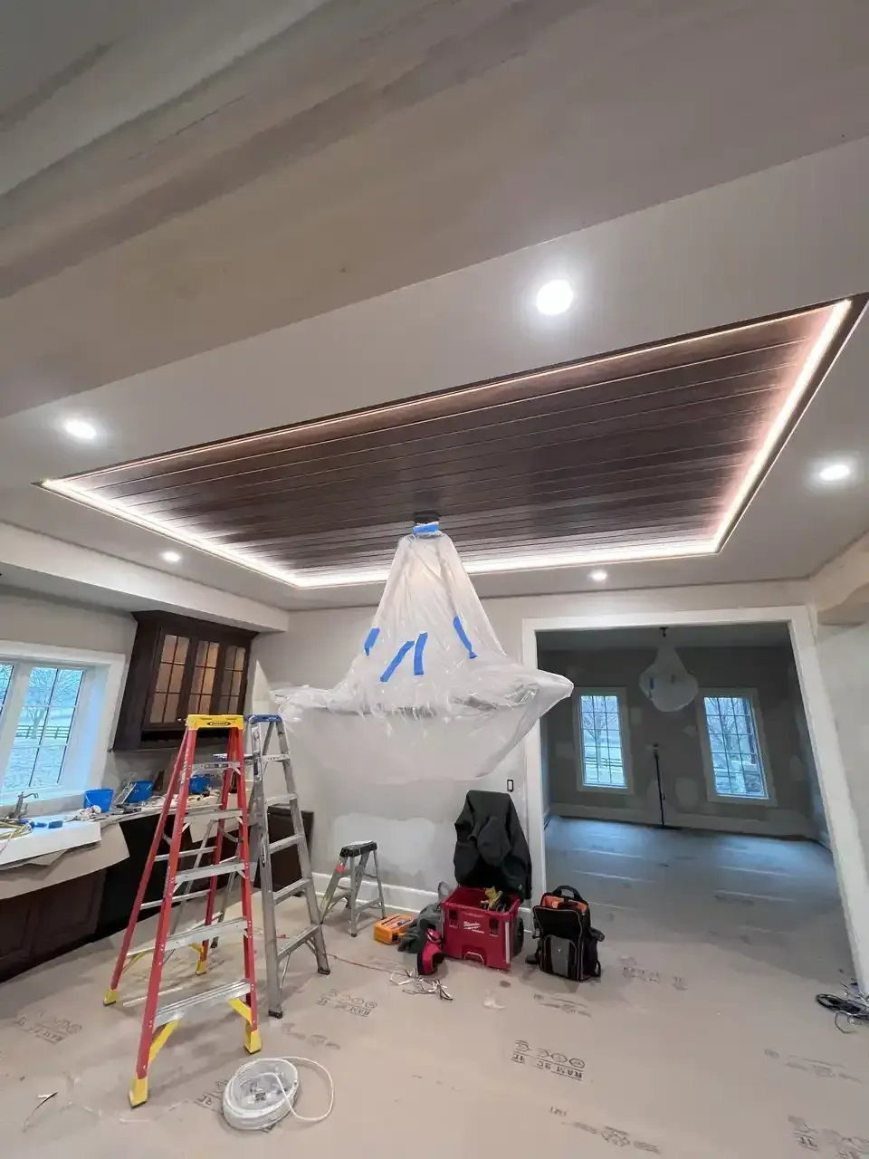 Room with recessed ceiling design, built-in lights, and partially covered chandelier, construction in progress.