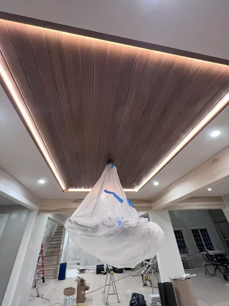 A ceiling with wood paneling and recessed lighting, with a chandelier wrapped in plastic below.