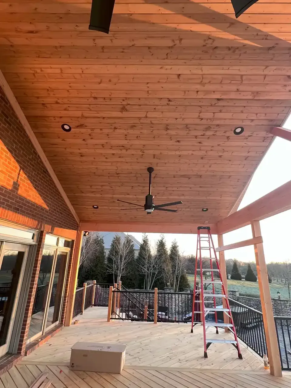 Outdoor deck with a wood plank ceiling, recessed lights, and a ceiling fan; a ladder leans against a beam.