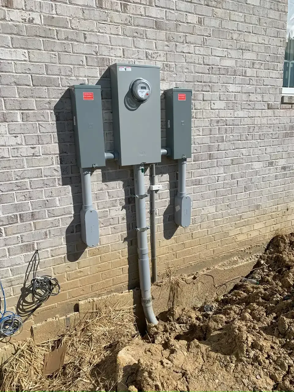 Electrical meter box and two breaker boxes mounted on a brick wall, with conduit emerging from the ground.