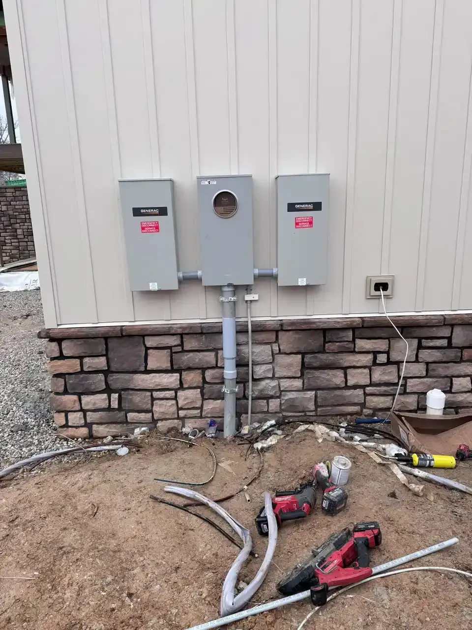 Electrical meter box and two breaker boxes mounted on a beige building with stone veneer base, tools in the foreground.