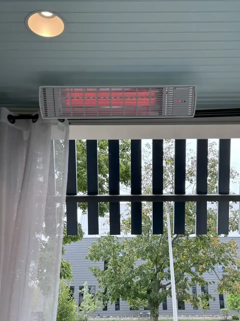 Ceiling-mounted infrared heater emitting red glow, mounted above outdoor seating area with blue ceiling and black canopy.