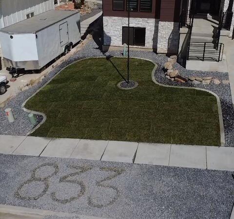 A small landscaped yard with green grass, a flagpole, stone edging, and the number 853 written on the sidewalk.