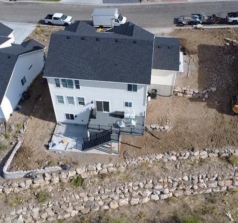 Aerial view of a two-story white house with a dark roof, front deck, and rocky shoreline nearby.