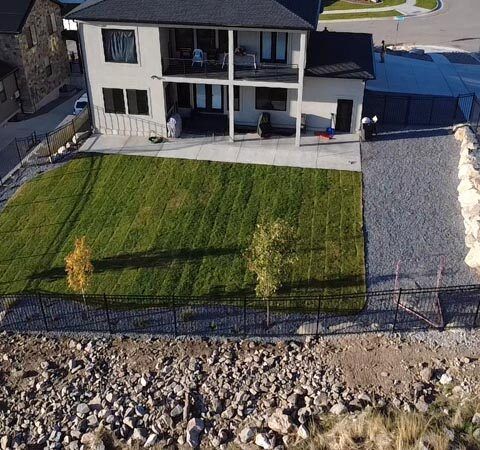 Aerial view of a modern house with a large green lawn, gravel driveway, and rocky shoreline in front.