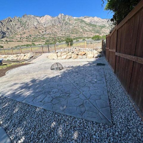 Stone patio with a fire pit, gravel border, wooden fence, and mountain view under a blue sky