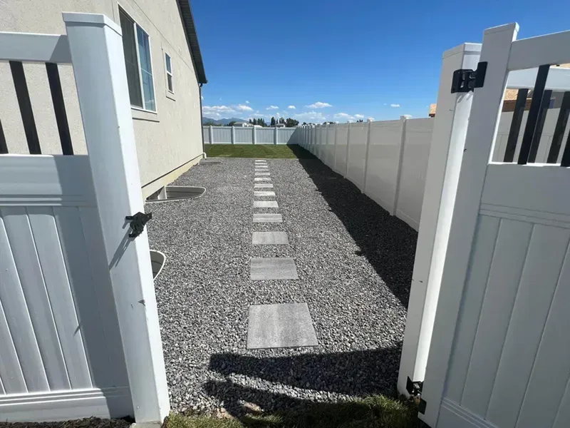 Narrow gravel walkway with stepping stones between white fences and houses under a bright blue sky