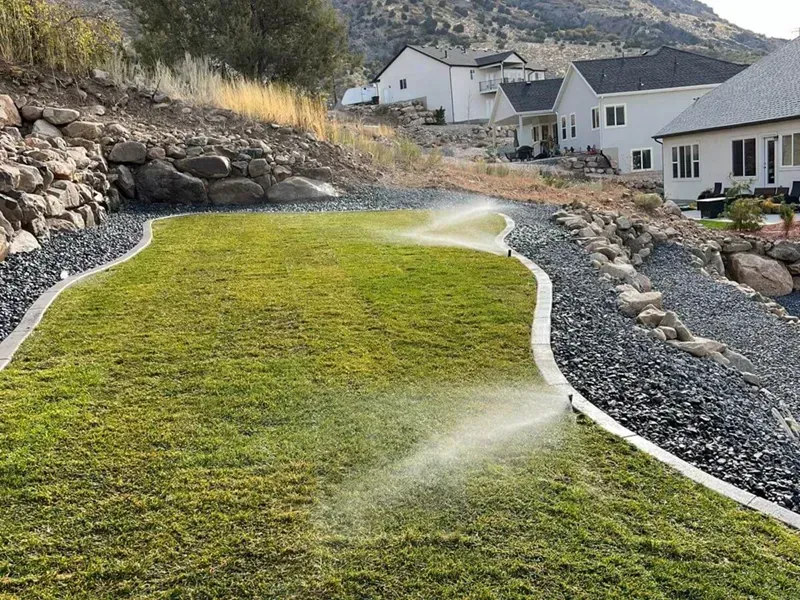 New sod lawn being watered by sprinklers beside a rocky slope and white houses