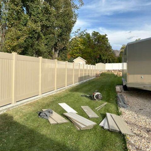 Row of beige fence panels along a grassy yard beside a parked trailer, with boards and tools laid out in front.