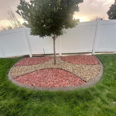 Circular rock garden with a small tree, red and tan stones, bordered by grass and a white fence.