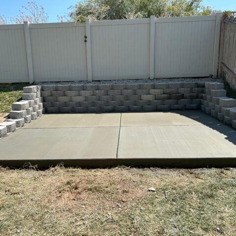 Concrete patio with a low cinder block retaining wall and white fence in a backyard