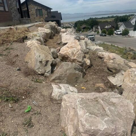 Rock-lined dirt slope beside houses overlooking a road and distant water under a hazy sky