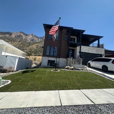 Modern house with an American flag, manicured lawn, mountain backdrop, and a white SUV in the driveway.