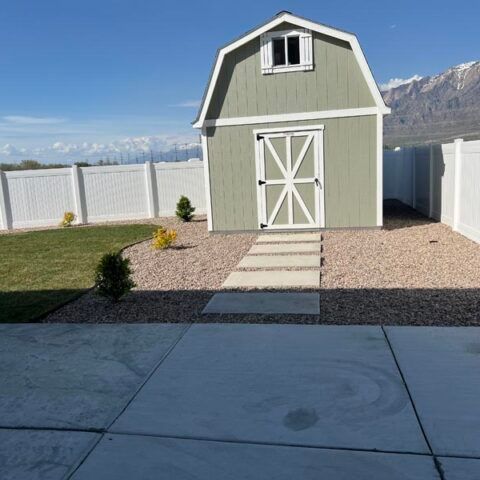 Green backyard shed with white trim, a small window, and a stone path in a fenced yard with mountains behind.