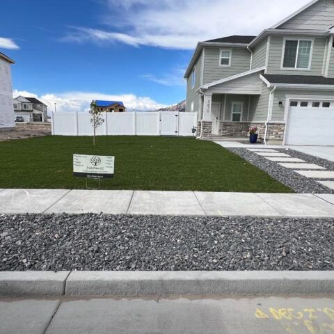 Suburban front yard with fresh green sod, white fence, and a gray house under a blue sky.
