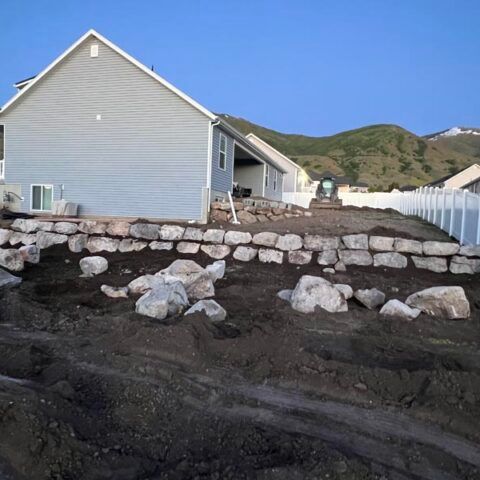 House beside rocky shoreline with dark debris in foreground and hills under a blue sky