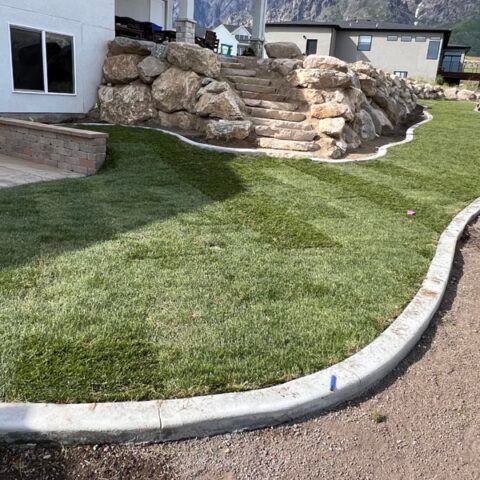 Landscaped yard with curved concrete edging, green grass, and a large decorative rock formation beside a house