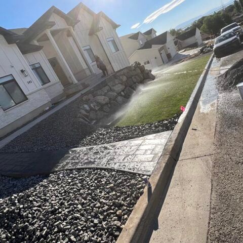 Suburban street with a landscaped rock drainage channel and water spraying across a sloped driveway.