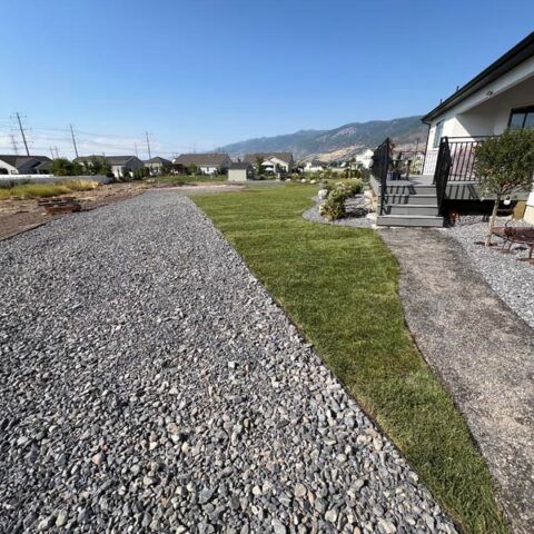 Gravel driveway beside a green lawn and house under a clear blue sky with mountains in the distance