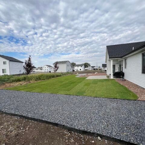Suburban backyard with green lawn, gravel path, and white houses under a cloudy sky