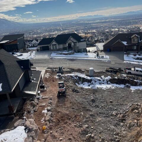 Hilly construction site with dirt, rocks, and houses overlooking a snowy valley and distant city.